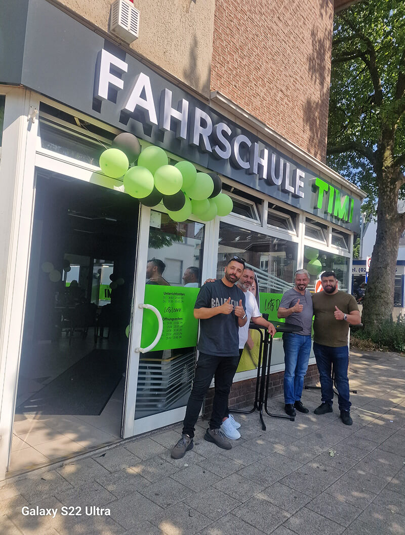 Eine lächelnde Gruppe von drei Personen vor einer Fahrschule mit grünen Luftballons.