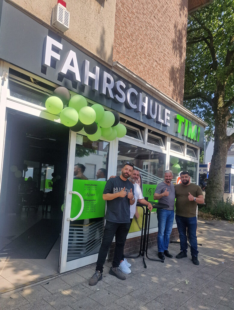Vier lächelnde Männer vor einer Fahrschule mit grünen Luftballons