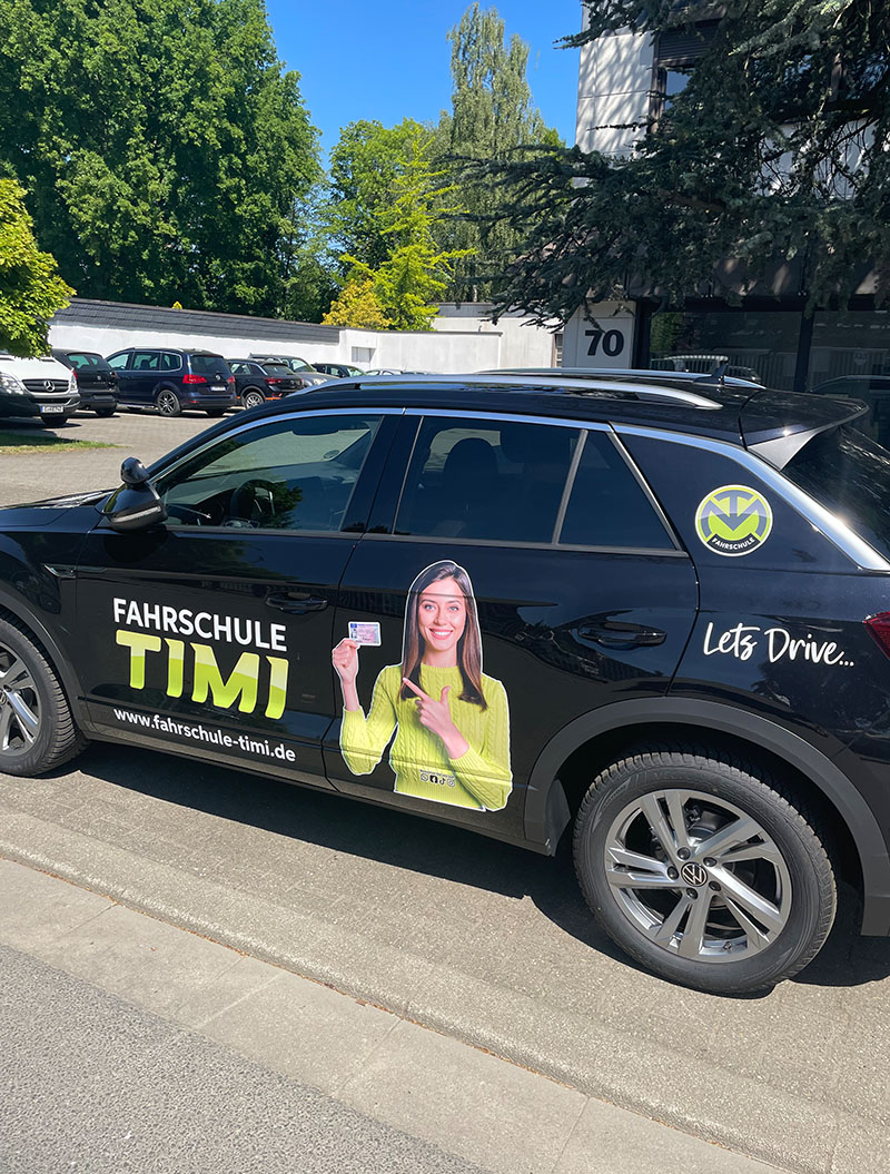 Eine schwarze Fahrschulauto mit einem Frauenselfie auf der Seite, lächelnd.