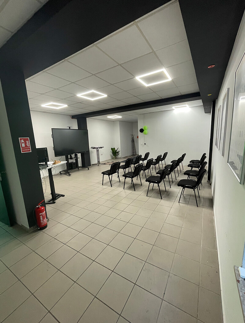 Ein leerer Raum mit Stuhlreihen, weißem Boden und einem schwarzen Bildschirm.