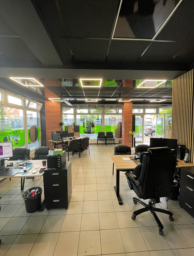 Ein modernes Büro mit schwarzen Decken und grünen Akzenten, leeren Schreibtischen und großen Fenstern, die viel Licht hereinlassen.